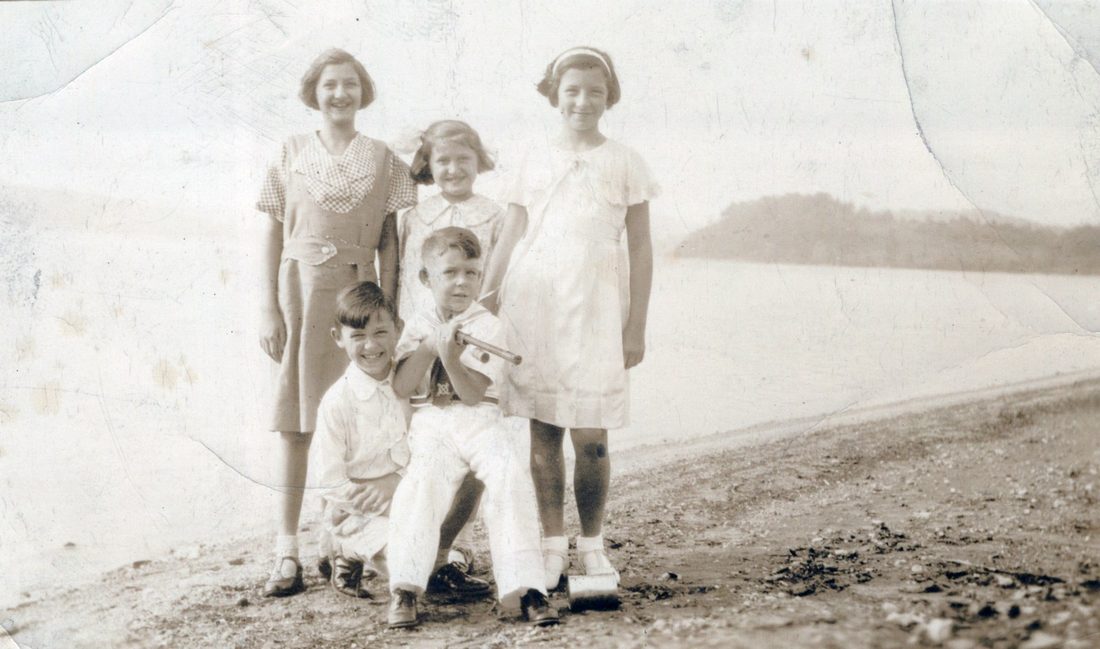 A faded sepia photograph of five children on a beach, three girls standing behind two small boys