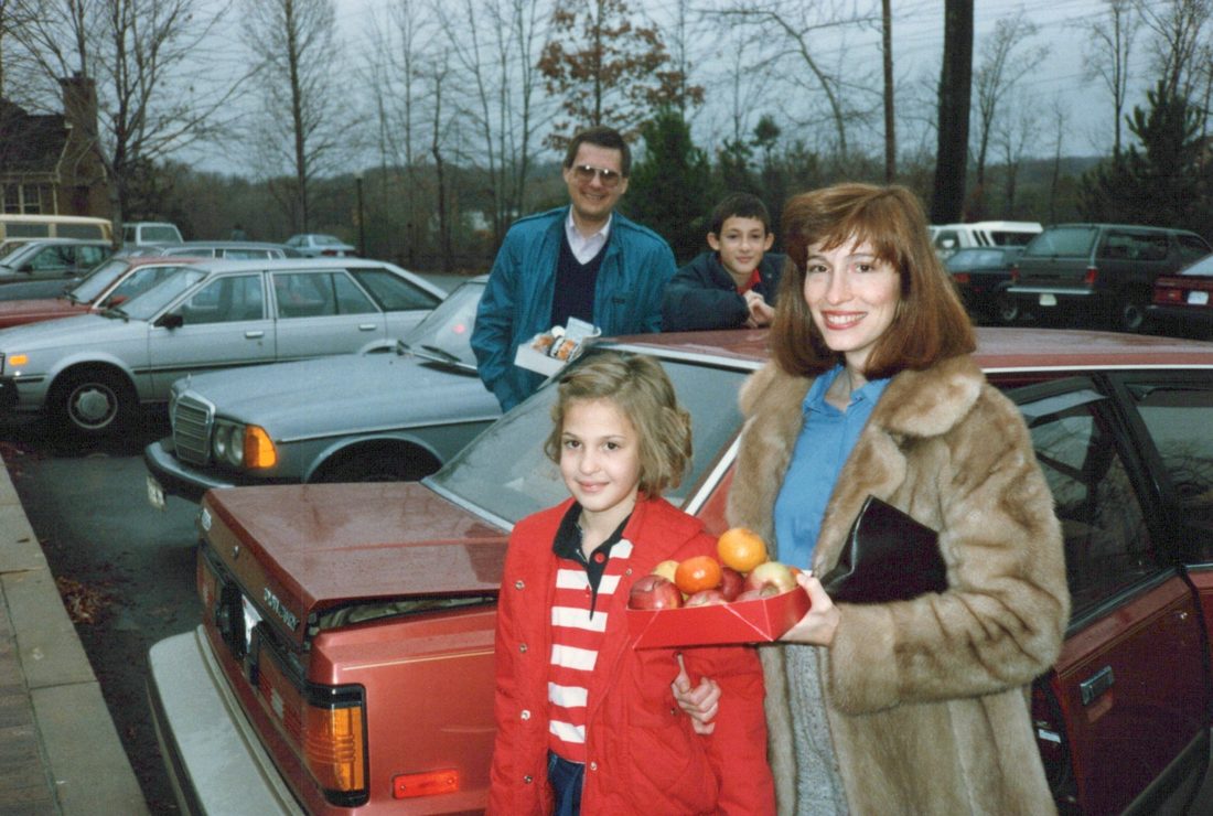 A colour photograph of a mother in a fur coat and her daughter in a red jacket holding a box of fruit beside a red car, a man and a boy behind them